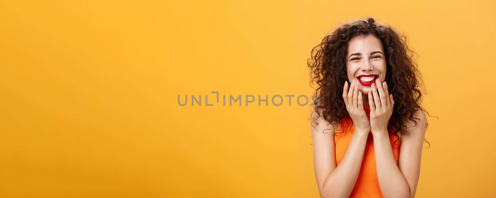 Waist-up shot of delighted charming and cute curly-haired caucasian female in cropped top being touched with sweet words smiling pleased holding palms on jaw-line surprised and happy over orange wall by Benzoix
