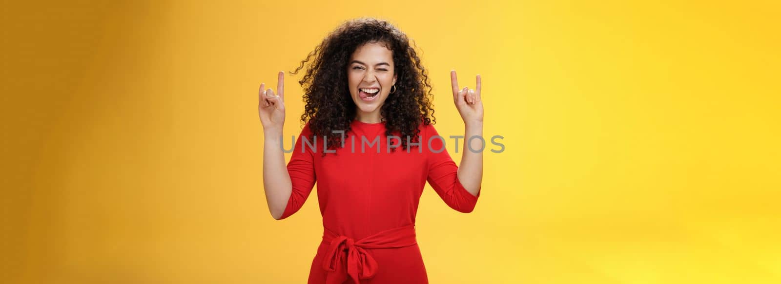 Cute excied and daring woman in 25s sticking out tongue as fooling around having fun on concert winking joyfully and showing rock-n-roll gesture feeling amused and alive over yellow background by Benzoix