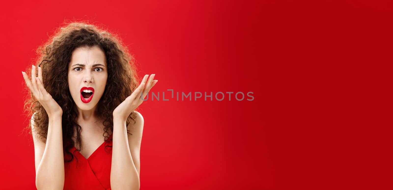Moody displeased and angry rich woman being dissatisfied with awful chip gift raising palms in frustration and clueless gesture arguing, shouting in outrage standing in red elegant dress. Copy space