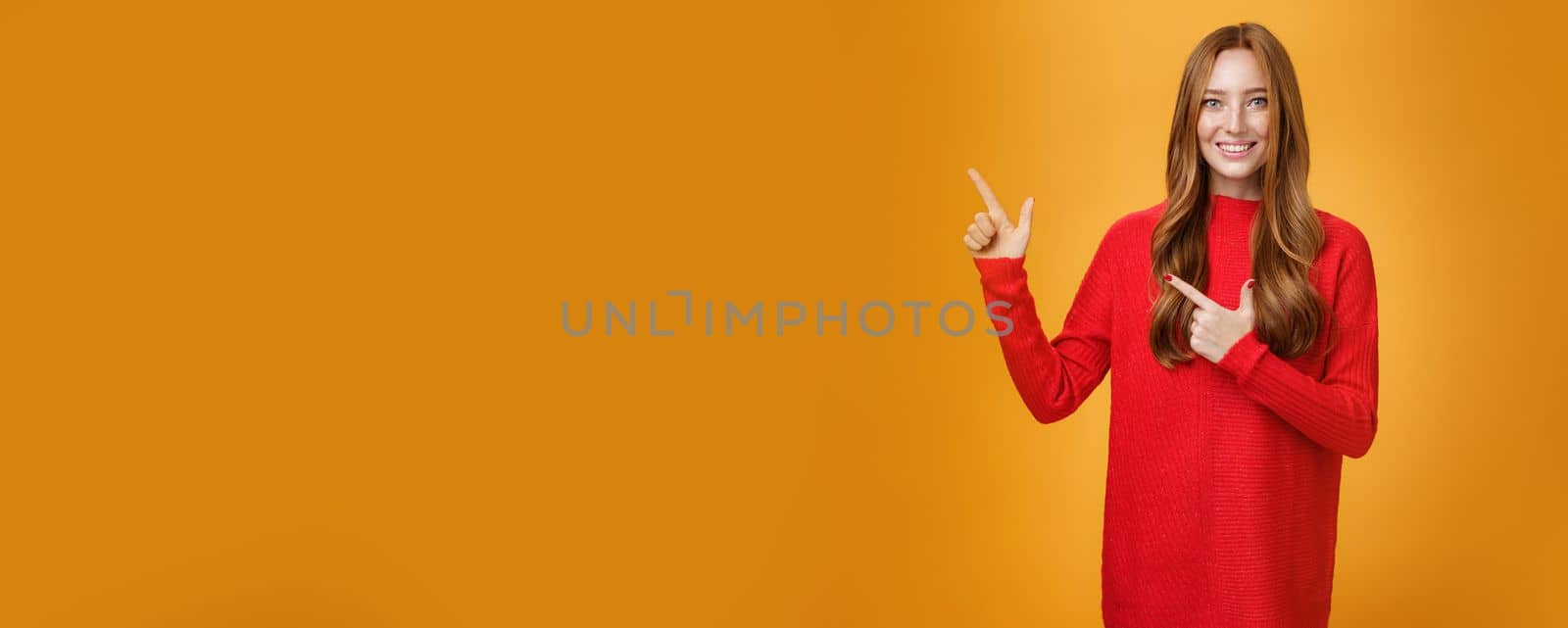 Assertive and helpful charming young female assitant in red knitted dress with ginger hair smiling friendly and pleasant as pointing at upper left corner, showing interesting thing over orange wall by Benzoix