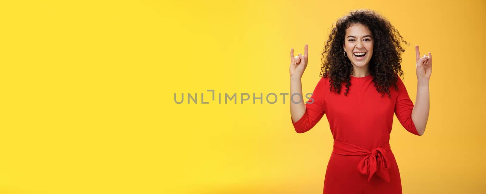 Rock n roll live. Charismatic excited young curly-haired woman in red dress feeling awesome as waiting concert of favorite band showing rock gestures with raised hands and smiling thrilled and happy.