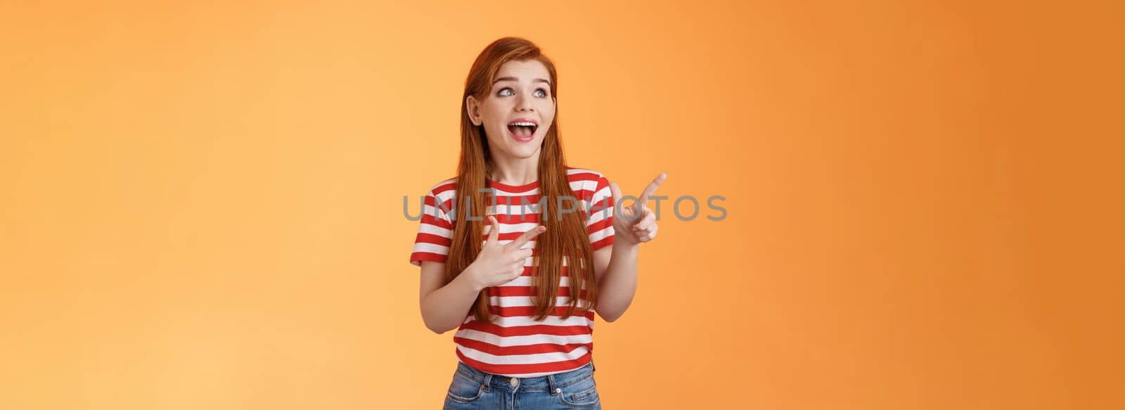 Enthusiastic cheerful redhead woman lively smiling laughing joyfully, enjoy watching copy space, look pointing upper left corner joyful, excited see awesome advertisement, orange background by Benzoix