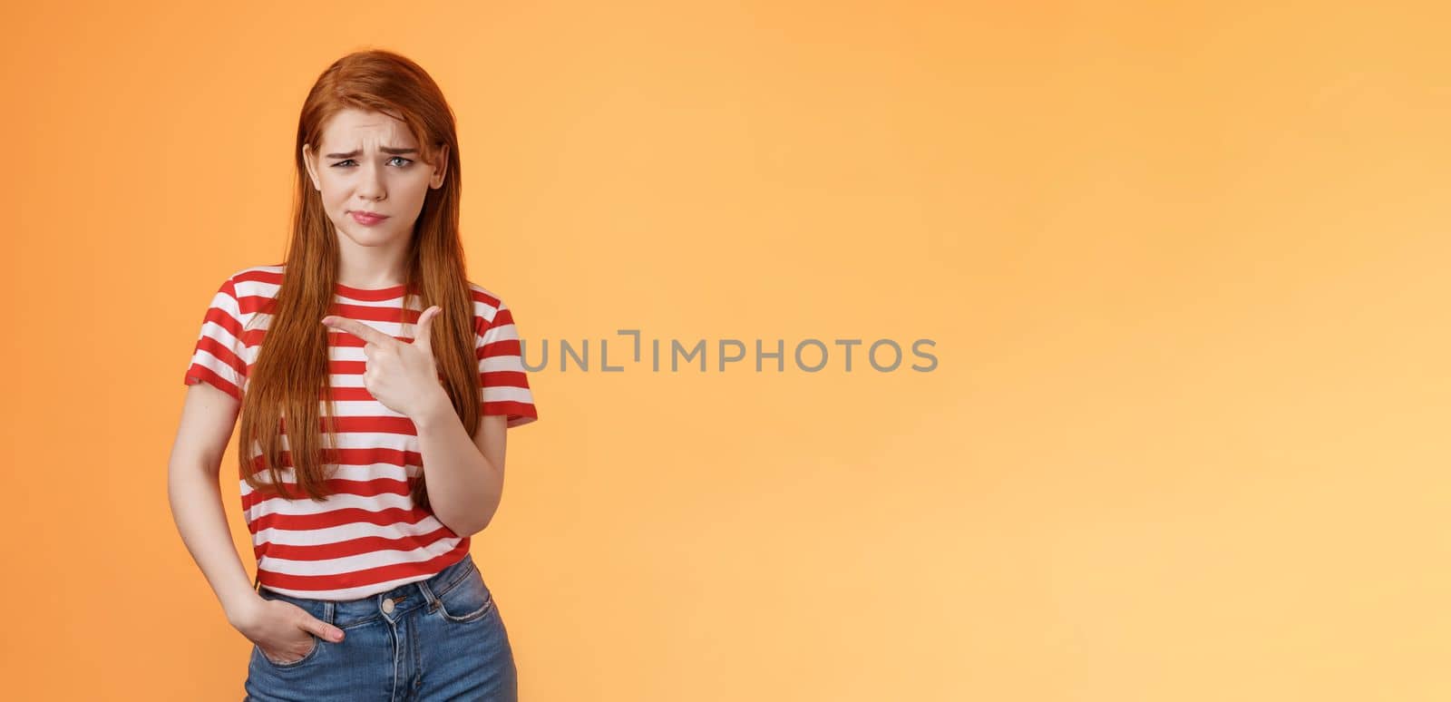 Upset uneasy cute ginger gitl long natural red hair, frowning smirking hesitant, complain, looking suspicious and doubtful point left, not sure if product really good, feel unsure, orange background.