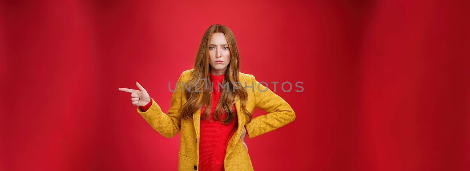 Explain yourself now. Portrait of upset gloomy and sad cute redhead girl waiting explanations asking question, pouting offended and pointing right with finger looking unhappy at camera over red wall.