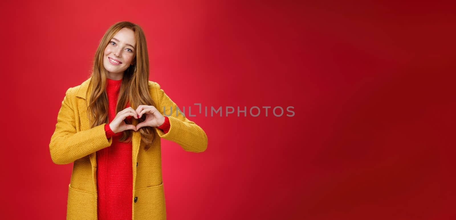 Love, romance and fall concept. Portrait of charming tender and gentle young redhead woman in yellow coat showing heart gesture making confession in sympathy, smiling cute at camera over red wall by Benzoix