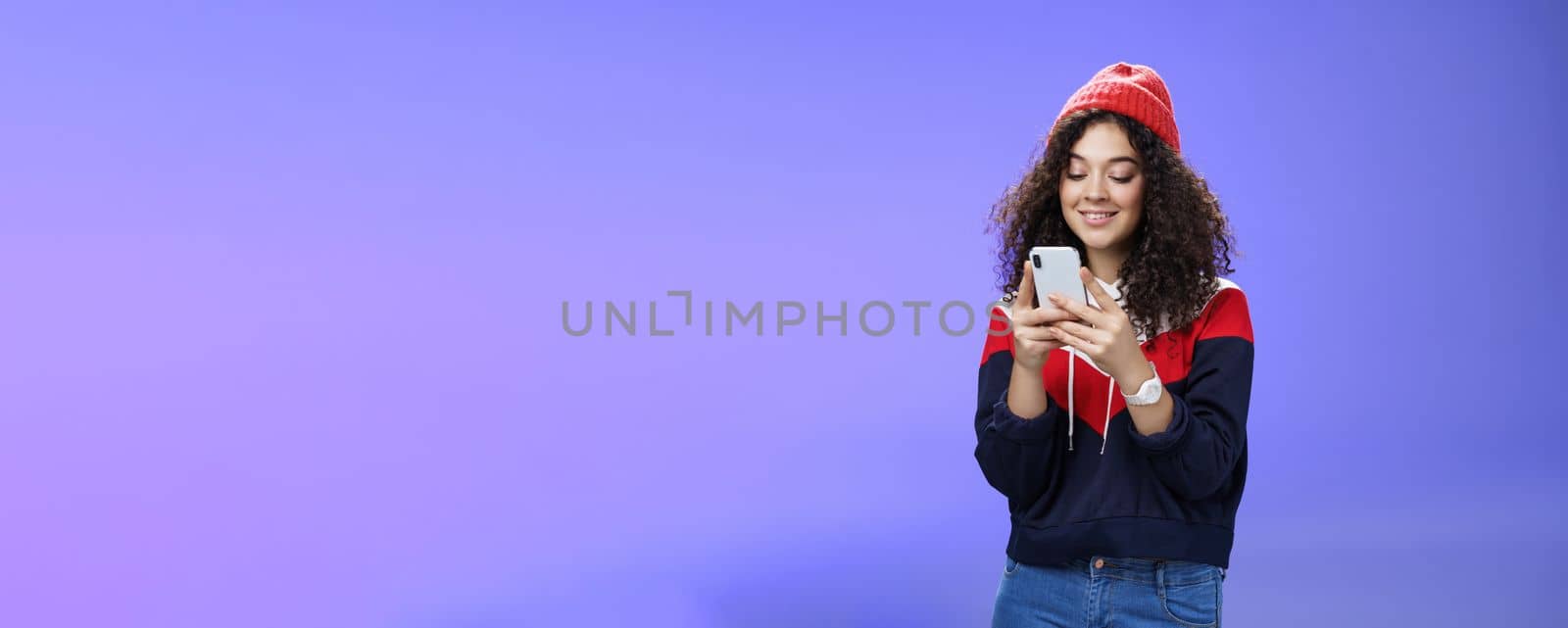 Woman searching in internet funny memes, holding mobile phone in hands smiling joyfully and cute at smartphone screen, typing message or browsing internet over blue background, wearing warm winter hat.