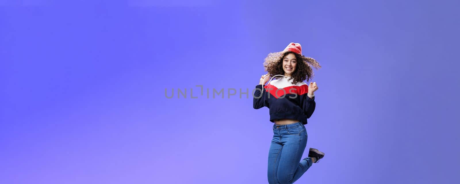 Delighted carefree and happy young woman with curly hair in warm beanie jumping joyfully having fun raising hands and smiling broadly posing over blue background in outdoor clothes by Benzoix