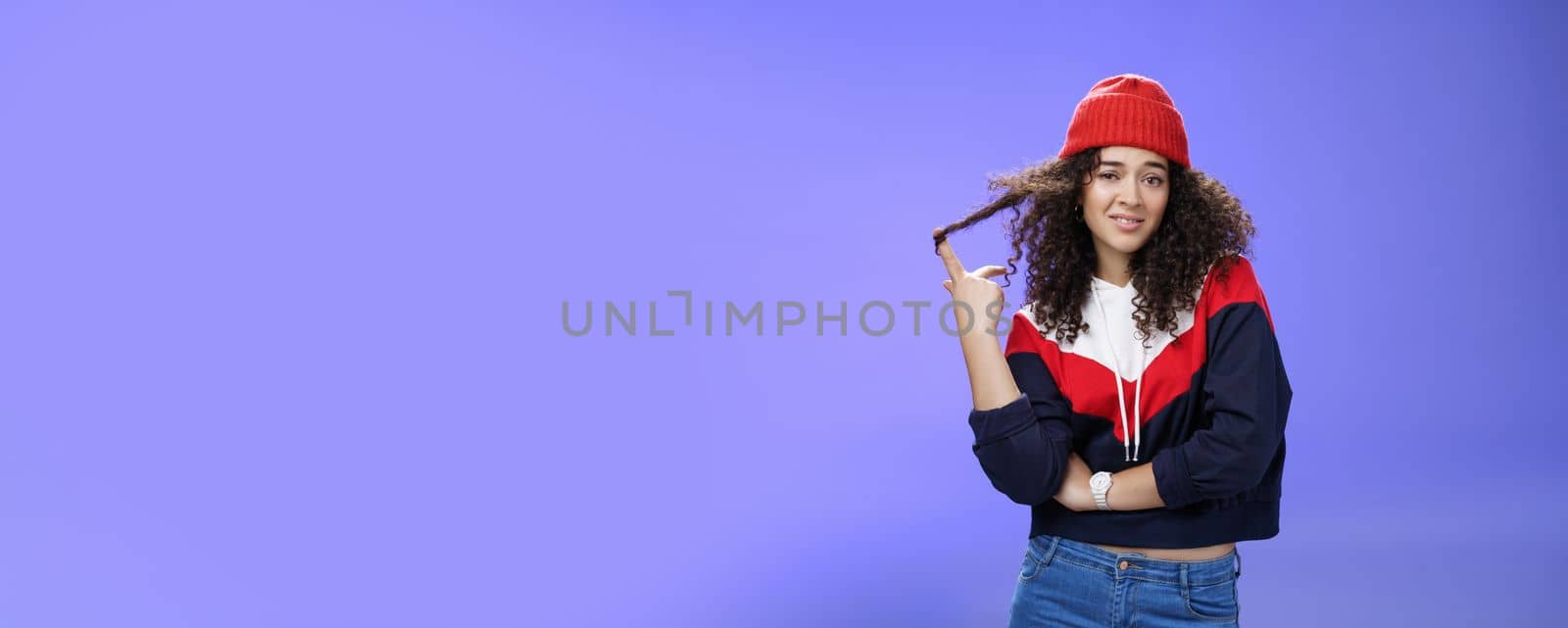 Stylish and snobbish arrogant curly-haired woman in warm beanie rolling curl on finger smirking and looking with contempt at camera, scorning person as being too cool over blue background.