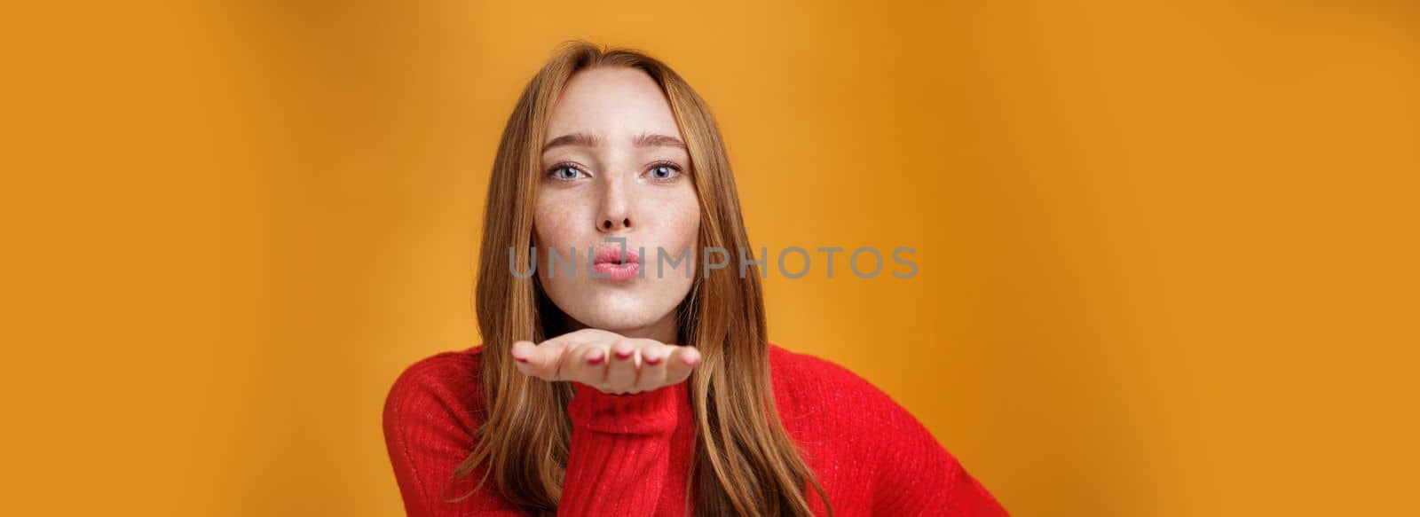 Close-up shot of sensual and attractive redhead female in red knitted outfit folding lips tilting forward as sending air kiss at camera giving mwah holding palm near mouth over orange wall by Benzoix