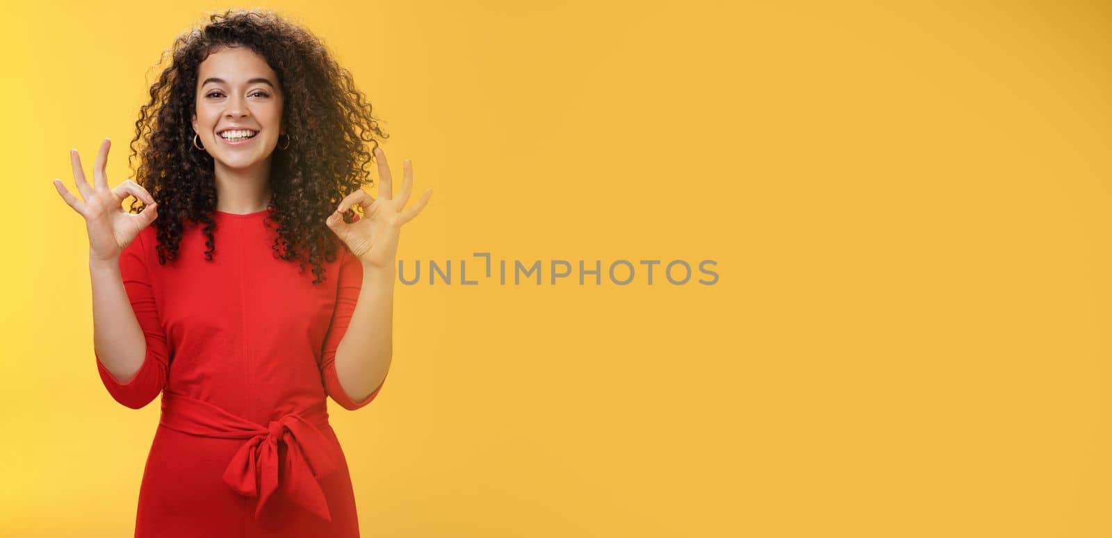 Girl pleased everything goes on plan and okay. Happy joyful charismatic woman with curly hair in red dress showing ok gesture in approval and like, satisfied with good work over yellow wall by Benzoix
