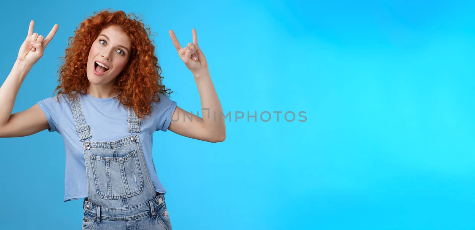 Lifestyle. Daring cool stylish awesome redhead cheerful curly-haired girl tilt head show tongue joyfully stare camera playful raise hands rock-n-roll heavy metal gesture having fun blue background.