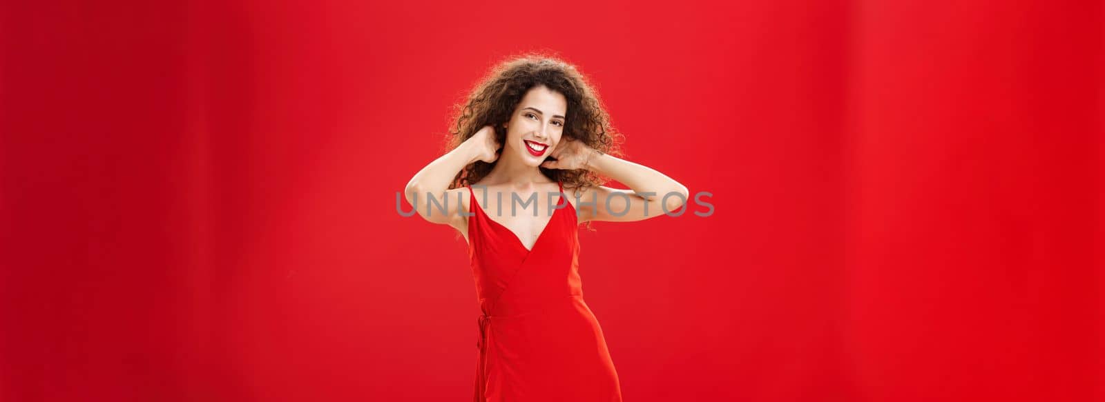 Stylish gentle caucasian girl with curly hairstyle and evening make-up touching strands of hair checking haircut and smiling broadly at camera posing in new elegant red dress over studio background. Copy space