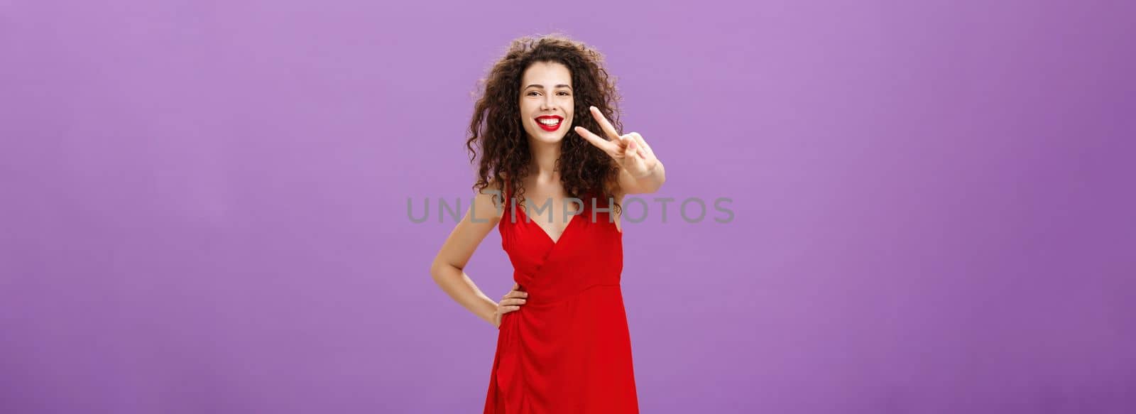 Studio shot of friendly gorgeous and stylish young european woman with cute curly hairstyle red lipstick and dress showing peace gesture at camera and smiling broadly over purple background. Emotions concept