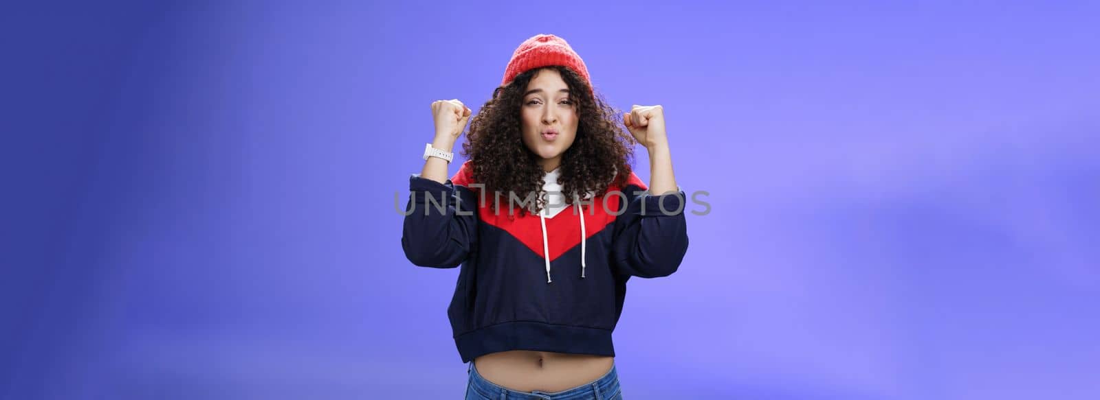 Charming young 20s girl with curly hair in warm beanie raising hands as belly showing under sweatshirt triumphing, celebrating awesome win folding lips from joy and excitement over blue background.