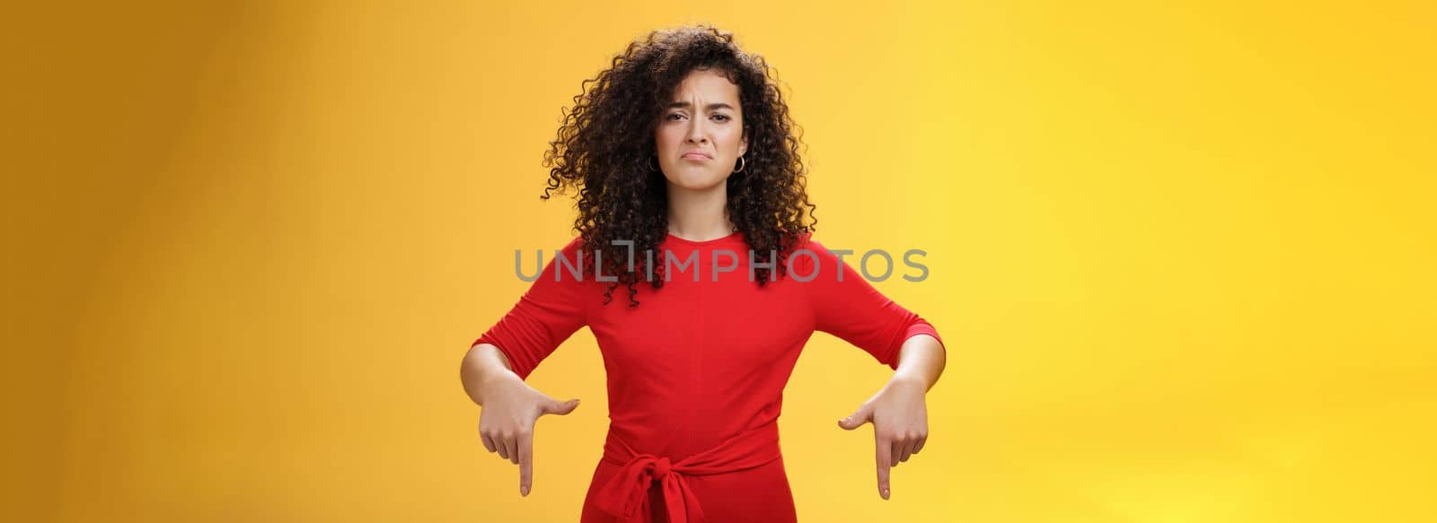 Gloomy disappointed cute silly curly-haired female in red dress frowning upset and making sad smile as pointing down with regret and unsatisfied expression, jealous or displeased over yellow wall.