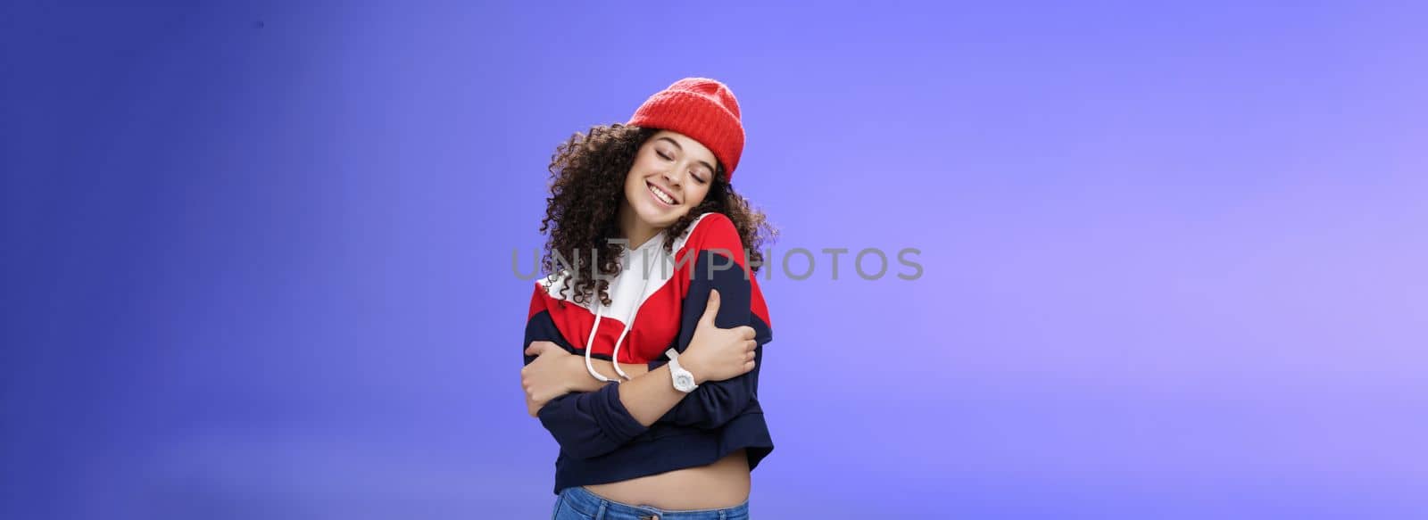 Lifestyle. Sweet tender and sensual curly-haired woman in red stylish beanie hugging herself with closed eyes and nostalgic smile leaning on shoulder feeling warmth and tenderness as recalling loving memories.