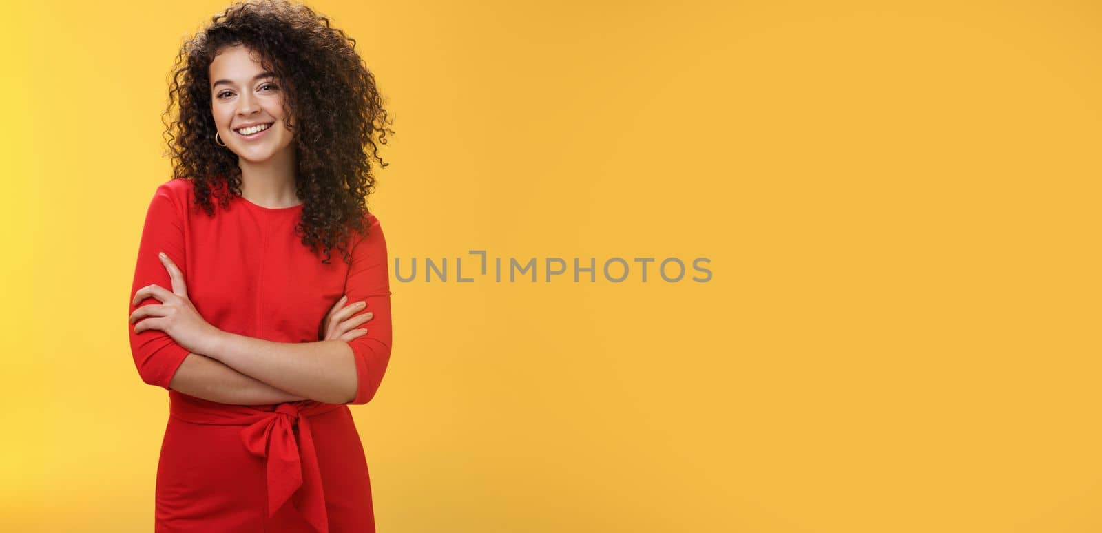 Confident charismatic young curly-haired woman in casual red dress holding hands crossed over chest in self-assured satisfied expression, smiling delighted posing over yellow background.