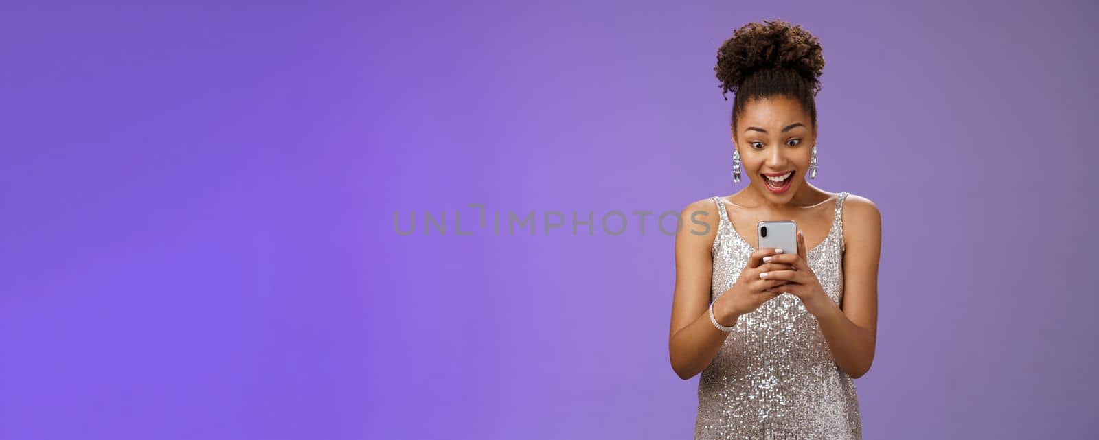 Good-looking amazed african-american woman in silver shiny dress smiling amused reading awesome news message smartphone look phone display astonished happy blue background by Benzoix
