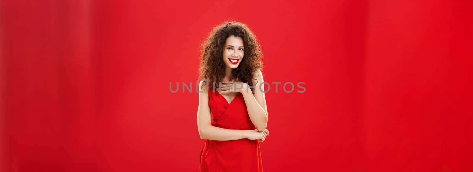 Woman receiving compliments attending luxury party in evening dress gently touching breast and flirting with sensual smile standing over red wall with curly hairstyle chuckling answering on guy flirt by Benzoix