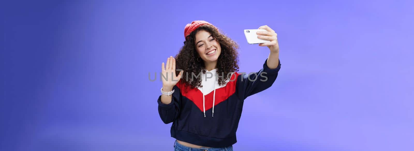 Studio shto of stylish cute female vlogger with curly hair in winter red hat smiling and waving hello at smartphone camera as recording video, making interesting content to post online over blue wall.