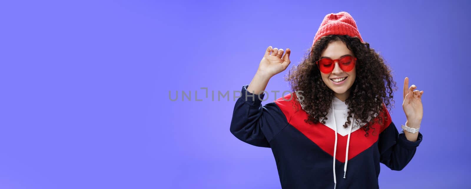 Girl feeling awesome having fun and enoying cool party, dancing with raised hands looking down smiling broadly wearing sunglasses and stylish beanie posing against blue background by Benzoix