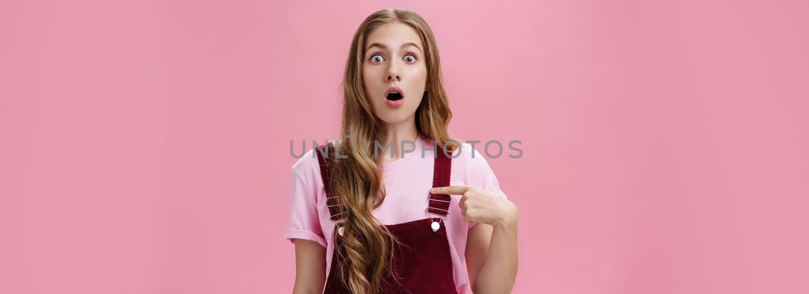 Shocked uncertain and confused charming young woman with tattoos and long natural wavy hair dropping jaw gasping and popping eyes from surprise pointing at herself with unsure expression. Body language concept