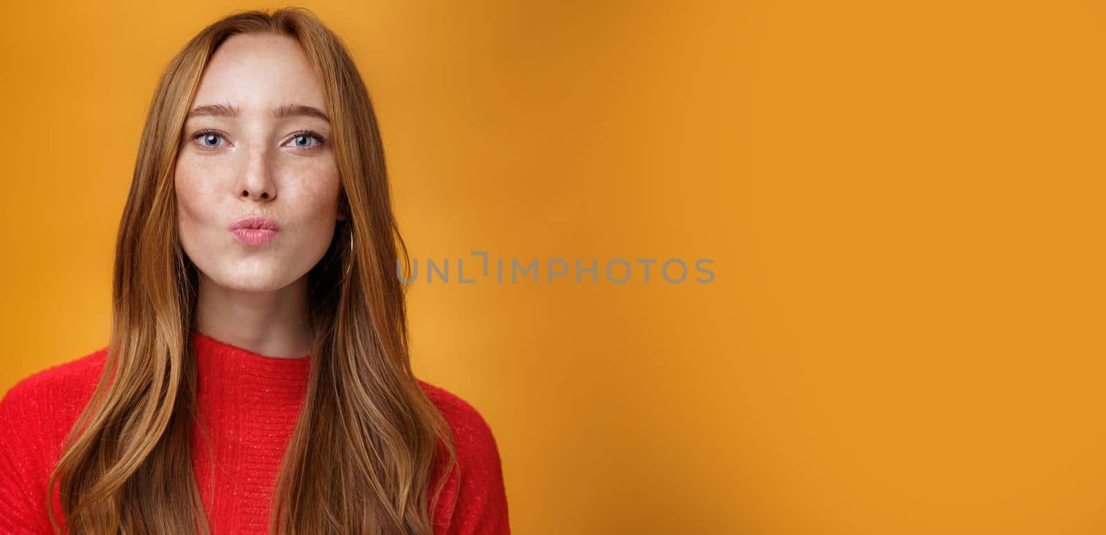 Close-up shot of flirty good-looking sensual redhead female in red knitted clothes folding lips in kiss and gazing at camera as giving mwah feleing romantic and pretty against orange background by Benzoix
