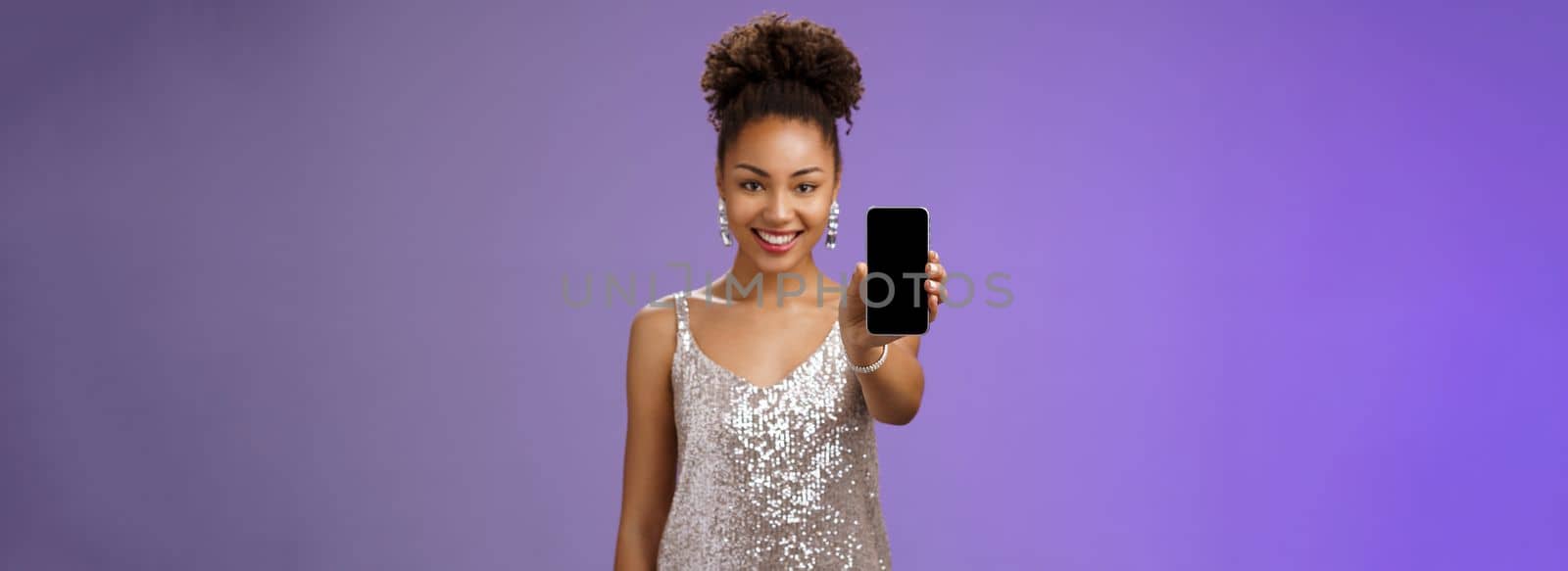 Charming attractive african-american woman in silver glittering stylish dress extend arm showing smartphone display presenting app promoting cool device standing pleased smiling blue background by Benzoix