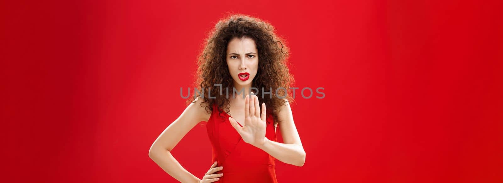 Hold on stop. Intense serious-looking displeased adult woman with curly hair in stylish red dress frowning raising palm in no gesture refusing giving negative answer, prohibiting come closer.