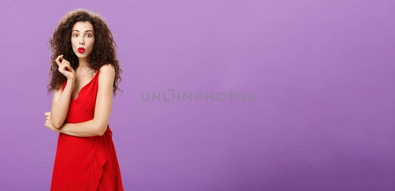 Intrigued chaming elegant lady with curly hairstyle. in red dress and lipstick folding lips curiously touching hair strand being amazed and impressed with rumors spreading around party over purple wall.