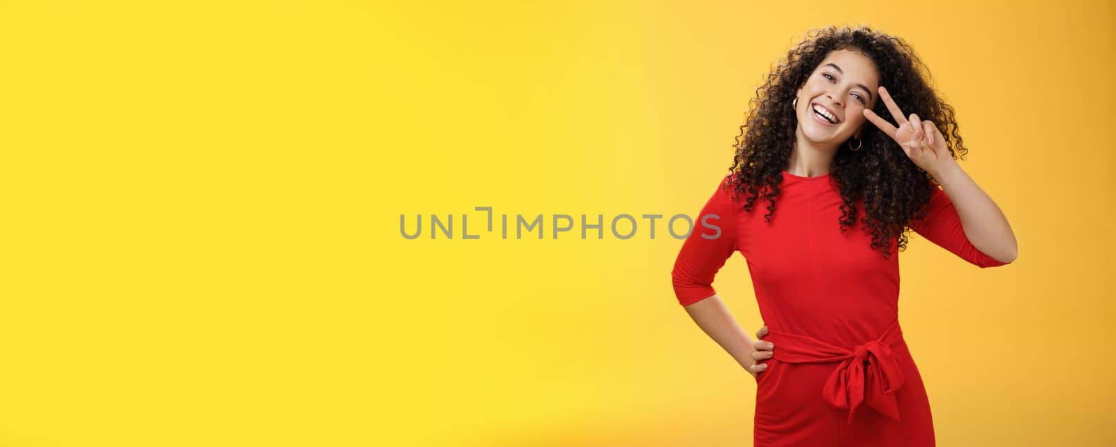 Waist-up shot of friendly tender and feminine cute woman with curly hairstyle tilting head and smiling, laughing and showing victory or peace gesture at camera over yellow background by Benzoix