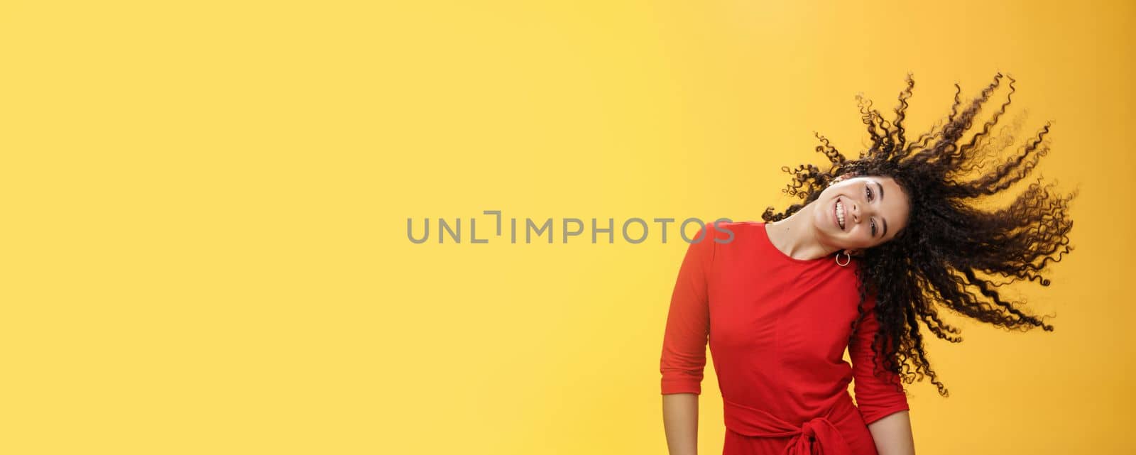 Girl getting carefree and wild waving hair and making sun with curls flying in air smiling broadly tilting head and having fun, dancing delighted and amused feeling playful against yellow background.