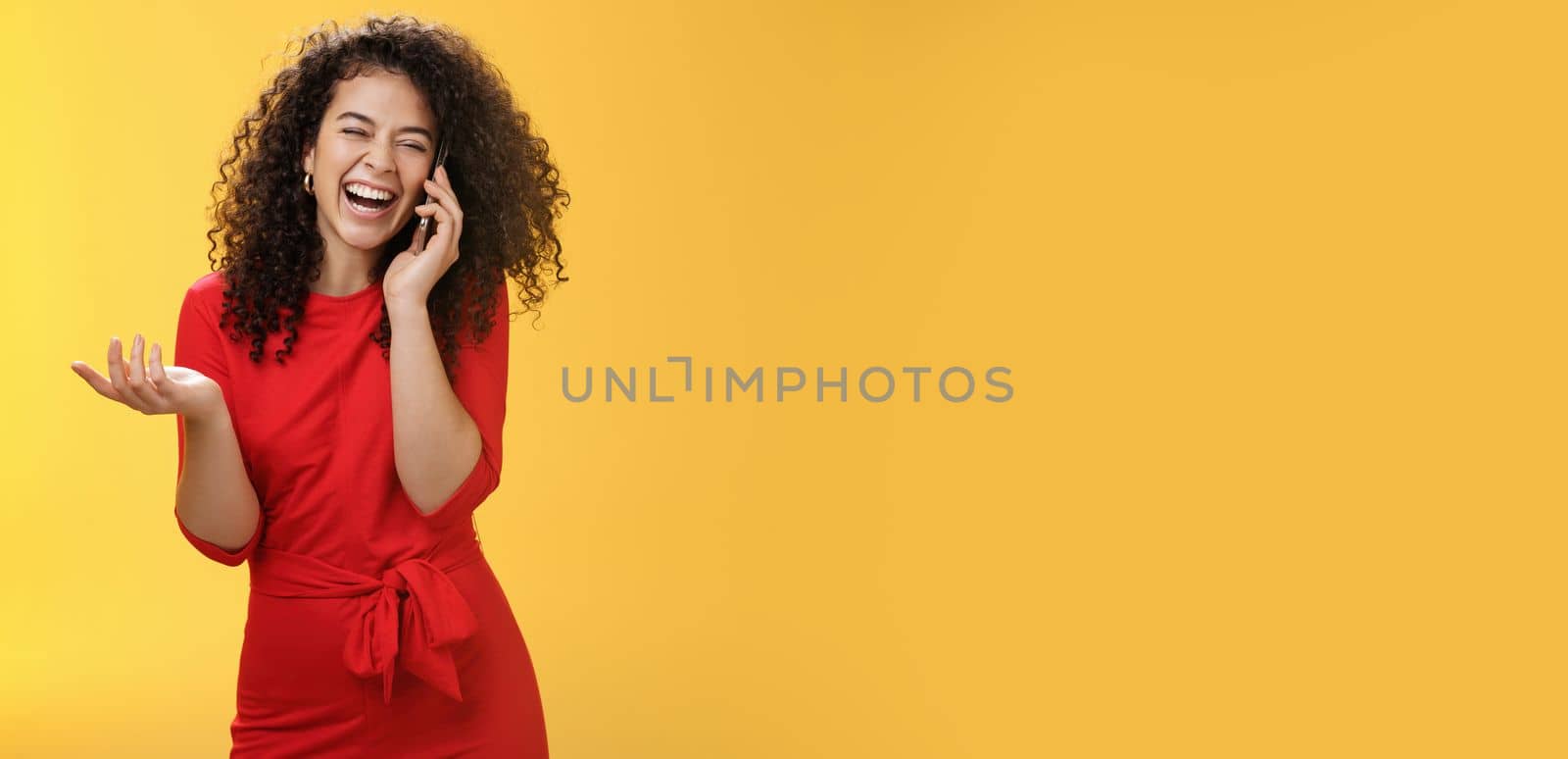 Portrait of joyful charming european female in red dress with curly hair laughing out loud as gossiping with friend via smartphone close eyes as giggling gesturing, hearing joke through mobile phone.