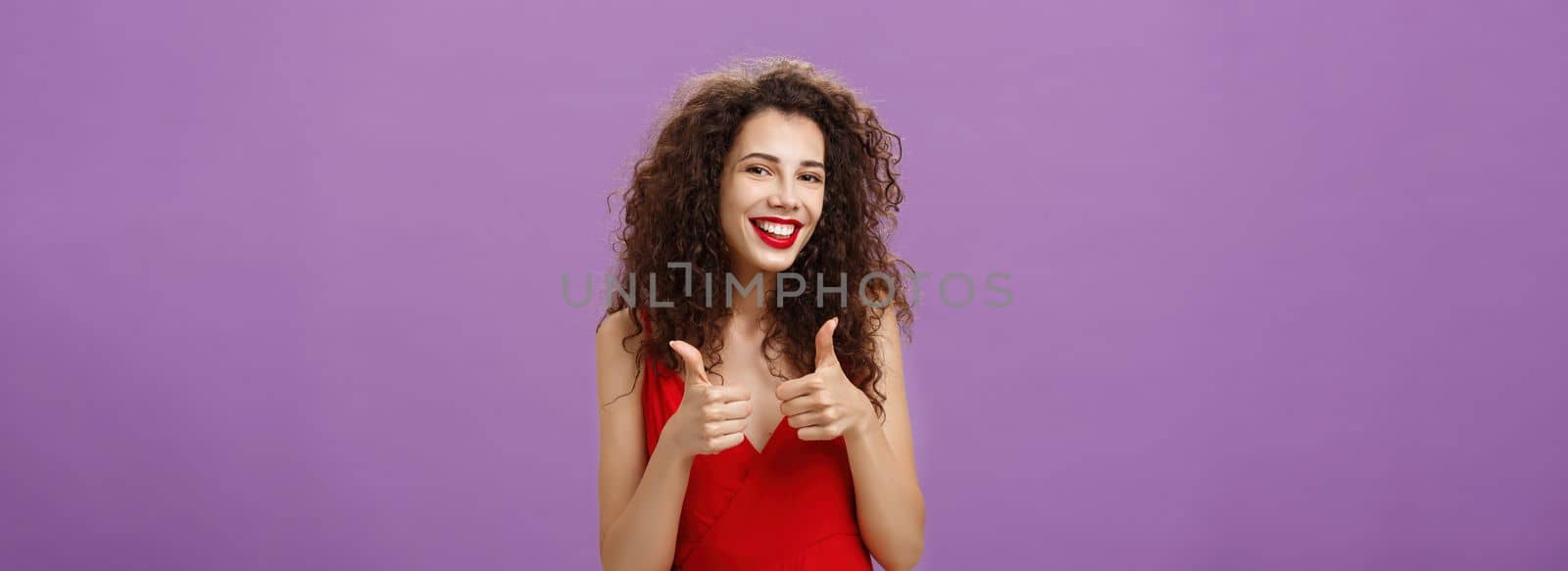 Supportive girlfriend giving thumbs up to decision of boyfriend move in. Friendly-looking tender charming woman with curly hairstyle in red dress giving approval and smiling over purple background.
