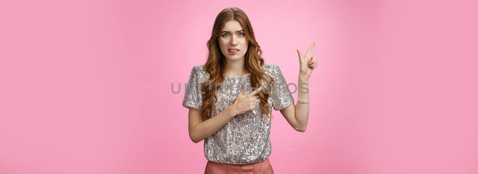 Dispelased insecure awkward young glamour rich girl dress-up party disappointed dislike place cringing aversion disgust pointing left questioned troubled, concerned standing pink background.