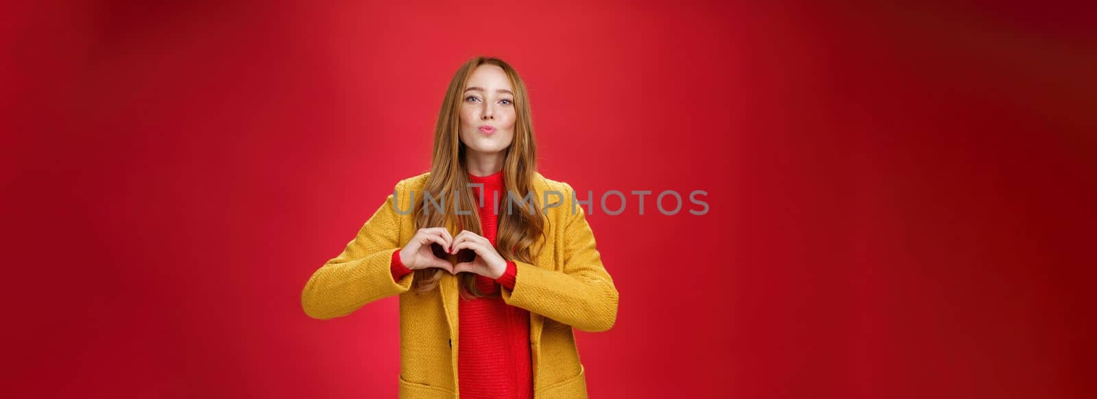 Love you all. Portrait of romantic and stylish good-looking flirty redhead female with freckles and blue eyes folding lips to give kiss showing heart gesture, confessing in sympathy over red wall by Benzoix