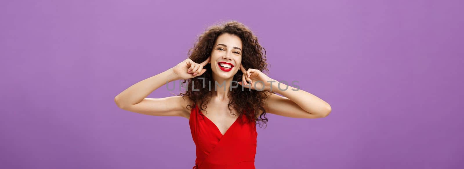 Prefer not listen and live life carefree. Joyful happy outgoing woman in stylish red evening dress closing ears with index fingers not hear about troubles posing delighted and joyful with broad smile.