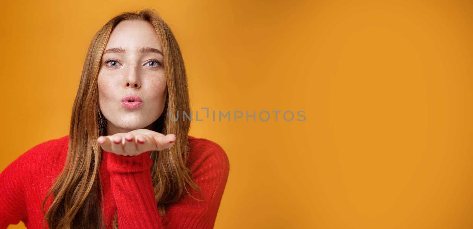 Close-up shot of sensual and attractive redhead female in red knitted outfit folding lips tilting forward as sending air kiss at camera giving mwah holding palm near mouth over orange wall by Benzoix