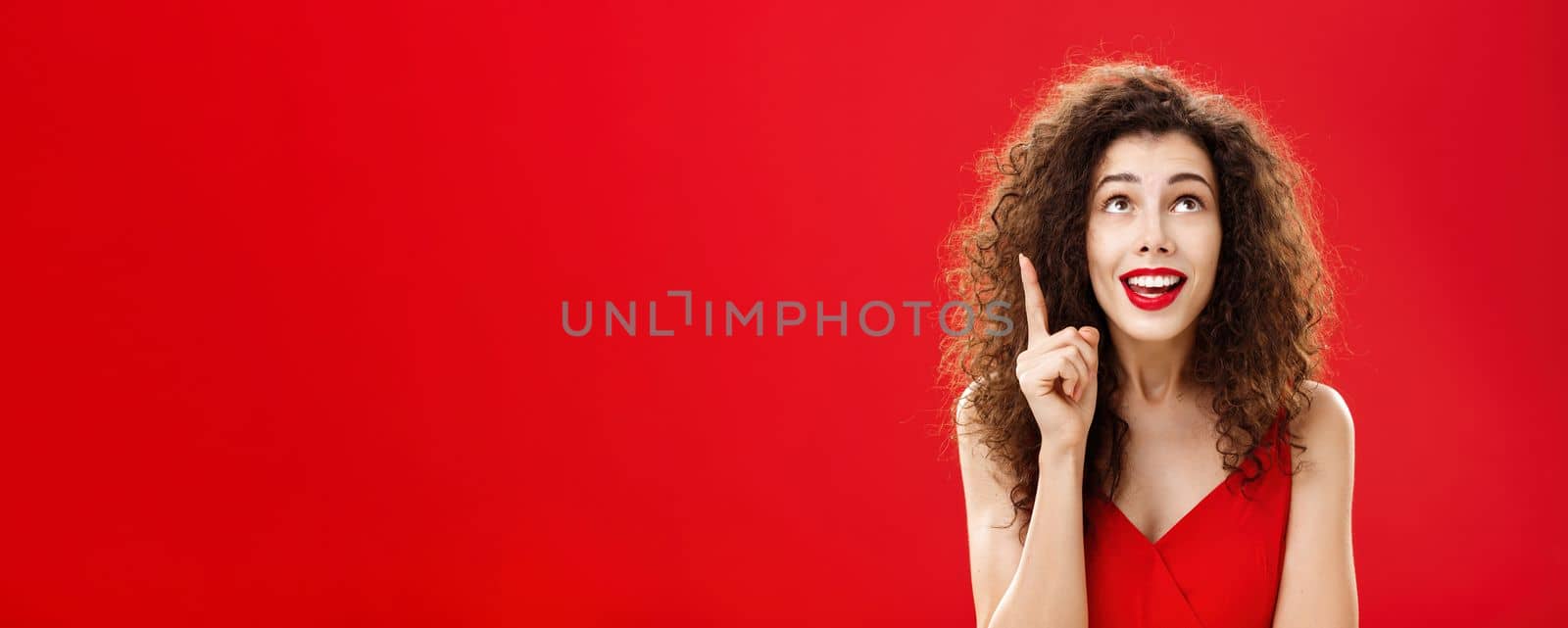 Portrait of delighted charming silly female with curly hairstyle. in dress raising index finger in eureka gesture smiling happily looking up having great plan or idea posing charismatic over red wall.
