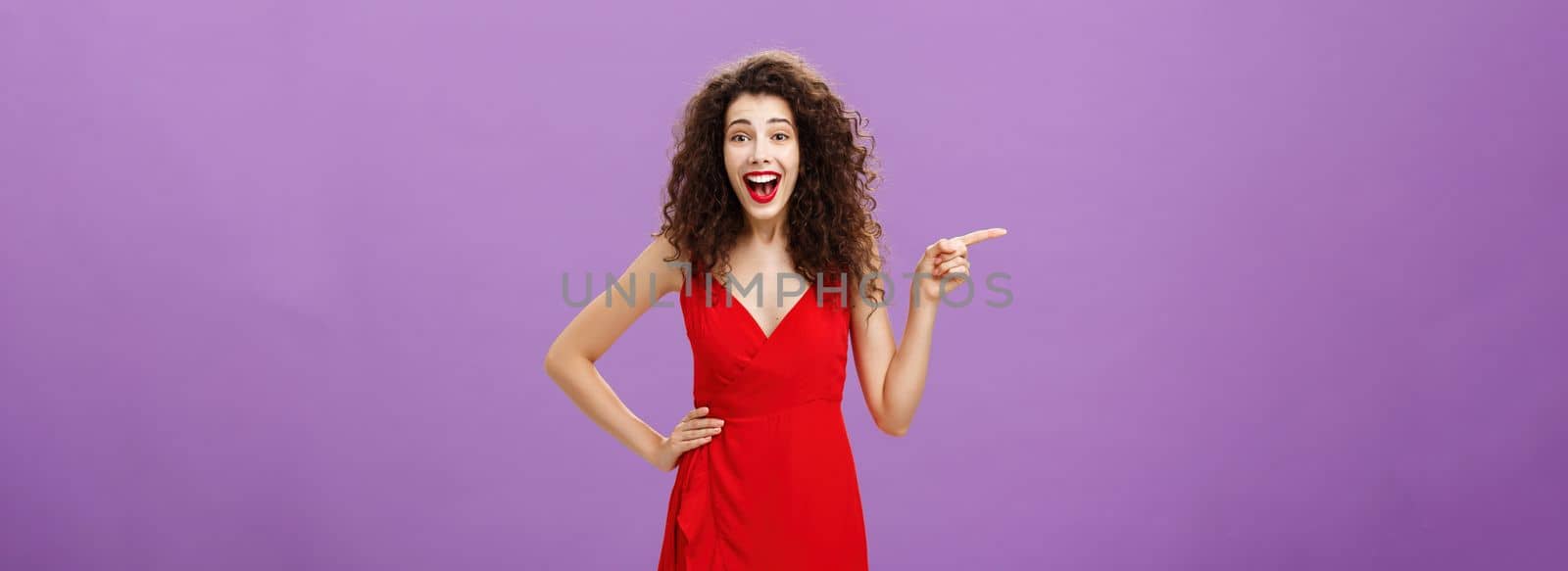 Portrait of entertained and amused joyful european woman. with curly hairstyle in red evening dress laughing from amusement and joy pointing left being entertained posing over purple background.