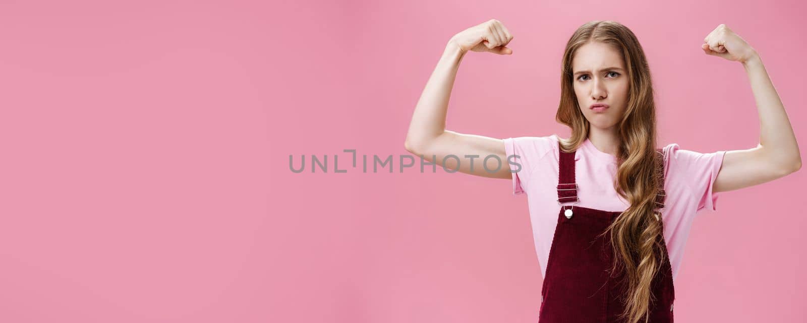 Girl shows she strong and independent. Serious-looking confident slender female raising hands to show muscles feeling self-assured in own power and female strengths posing against pink wall. Copy space