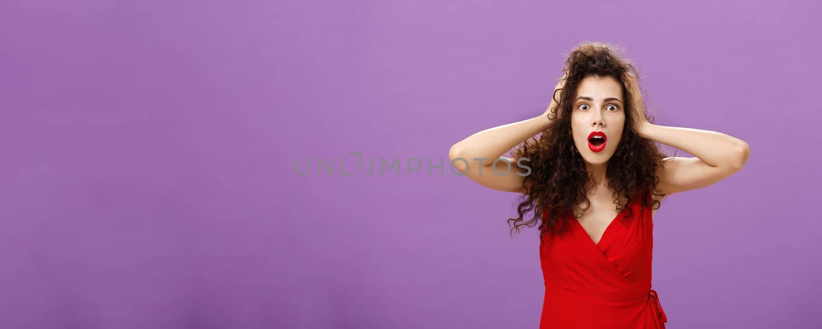 You do not say. Portrait of shocked and impressed speechless woman with curly haircut in luxurious evening dress opening mouth from amazement and shock holding hands on head stunned over puple wall.