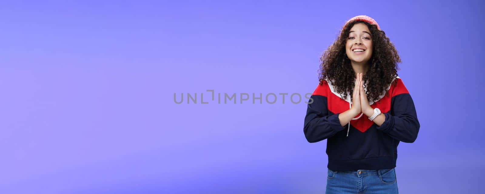 Charming excited and friendly-looking cute female friend with curly hairstyle in outdoor hat and sweatshirt holding hands in pray and smiling with angel expression as hoping friend help over blue wall by Benzoix