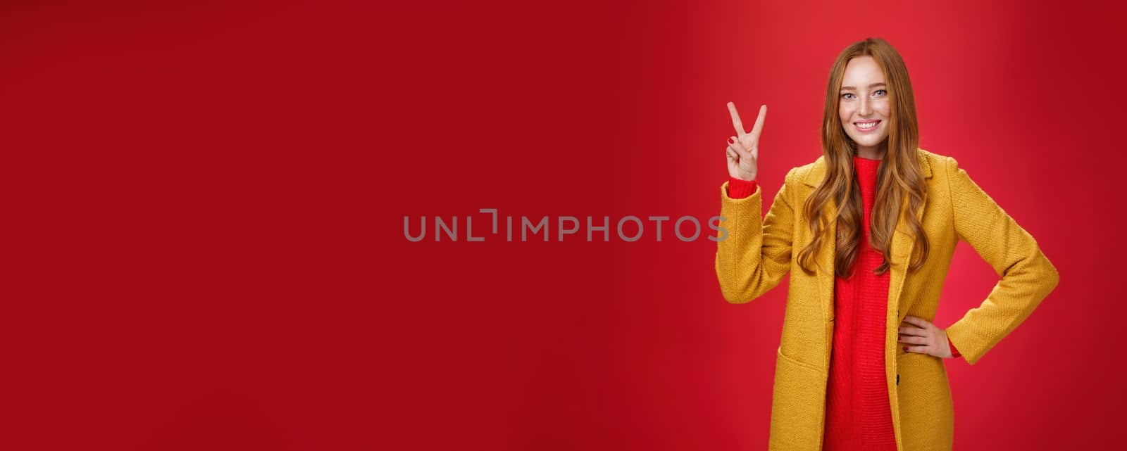 Portrait of happy playful and cute redhead woman with freckles in yellow coat holding hand on waist confident and self-assured showing peace or two sign delighted, beating weather conditions. Copy space