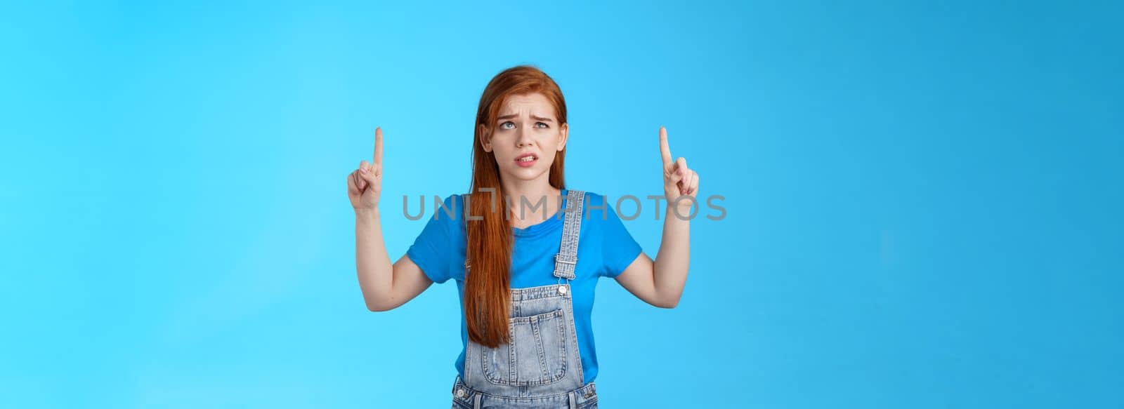 Uneasy worried cute redhead female coworker feel unsure insecure, grimacing displeased intense worried expression, look pointing up, top copy space, have hesitations, stand blue background.