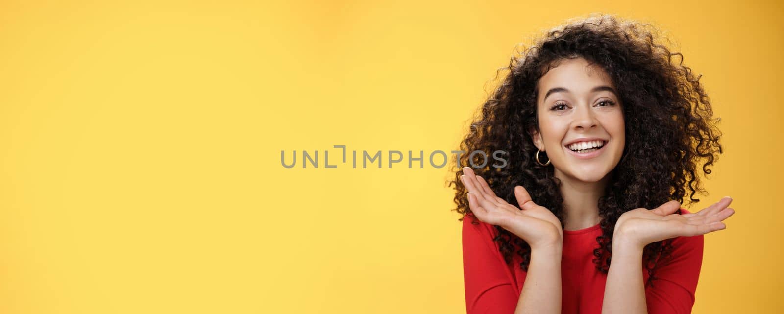 Lifestyle. Close-up shot of happy kind and tender pretty caucasian female student with curly hair and perfect skin smiling delighted holding palms spread near face having fun over yellow background.