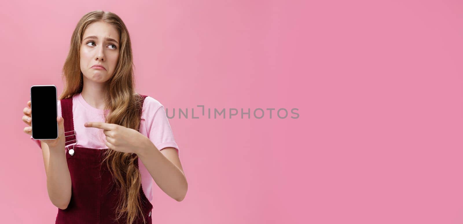 Indoor shot of gloomy displeased and disappointed cute young girl holding smartphone pointing at cellphone screen making upset grimace as if being bothered and dissatisfied against pink wall. Emotions and technology concept