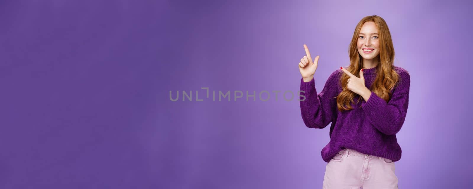 Lifestyle. Charming assertive female shop assistant pointing at upper left corner to promote cool product smiling broadly feeling joyful and excited expressing friendly attitude as posing in purple sweater.