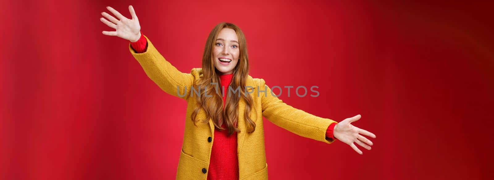 Hey come and hug me friend. Charming playful and emotional redhead female extanding palms forward to cuddle and greet, giving warm welcome smiling broadly posing in yellow coat over red wall.