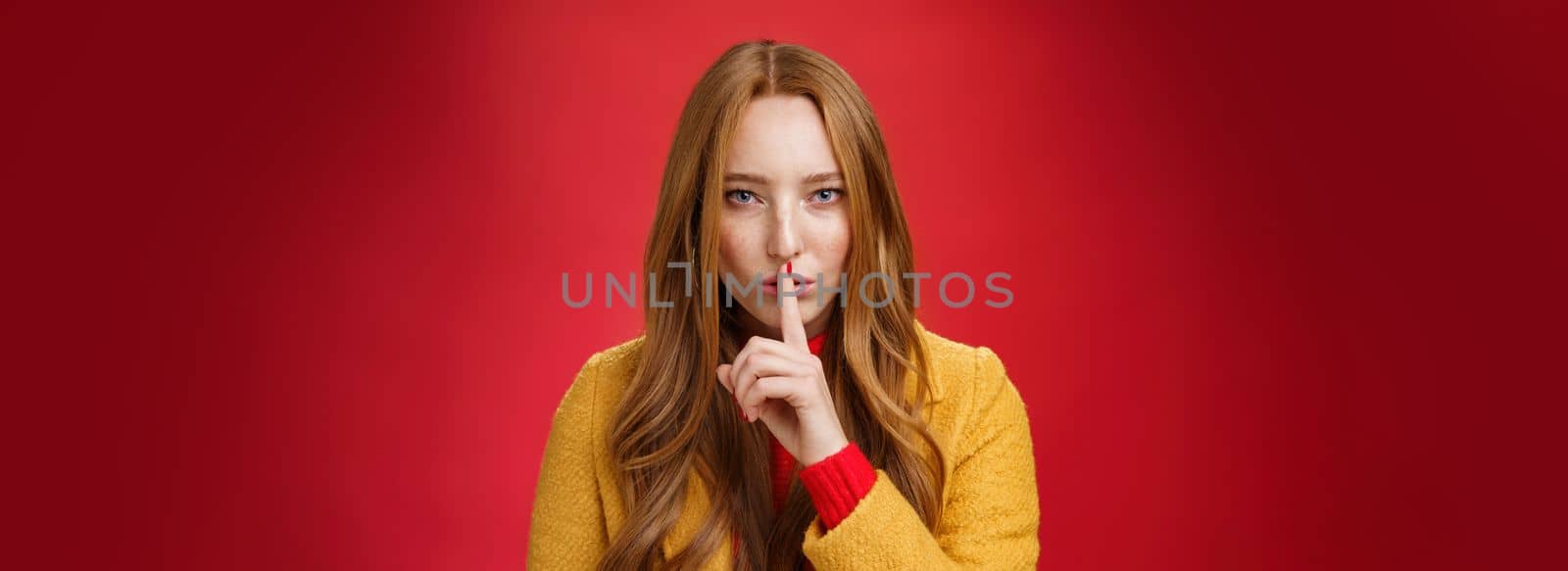 Shh keep out secret safe. Portrait of sensual attractive and daring redhead female in yellow coat showing shush gesture with index finger on lips, squinting looking determined and sassy over red wall.